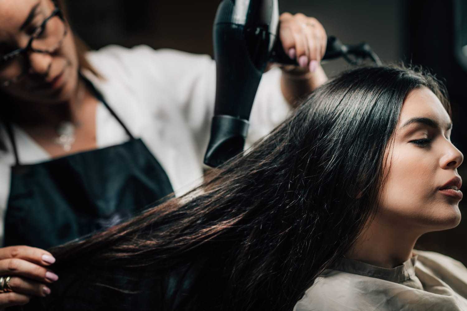 Hair stylist drying client's long hair with a blow dryer in a salon setting.